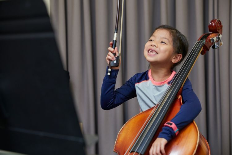 A girl with black hair wears a blue and pink top. She sits with a cello in one hand and holds a bow in the other. She has a big smile across her face.